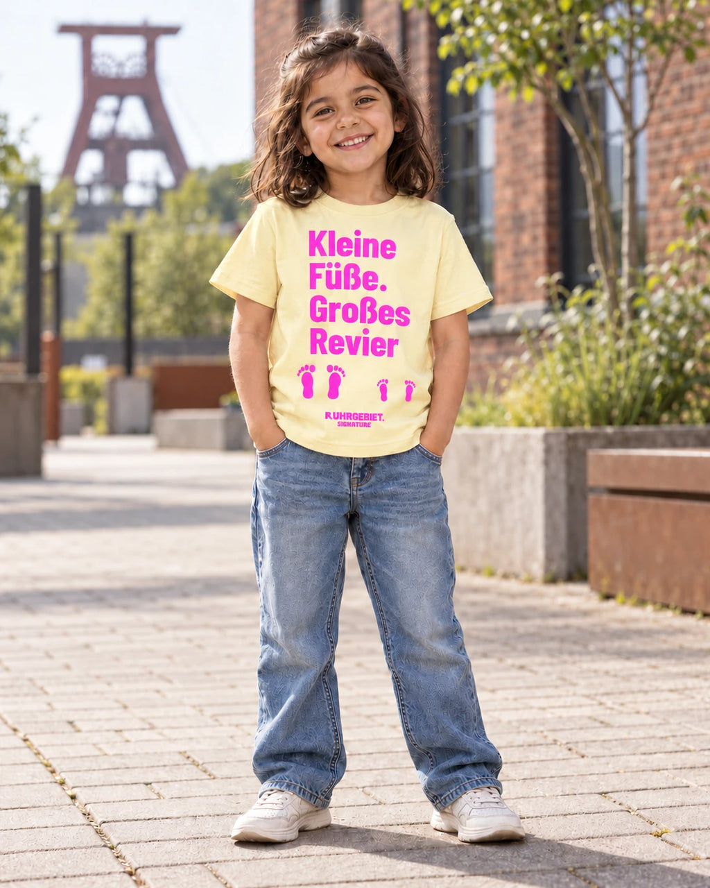 Fiktives Kind im buttergelben T-Shirt mit pinkem Motiv „Kleine Füße. Großes Revier“ bei Tageslicht. KI-Produktillustration.