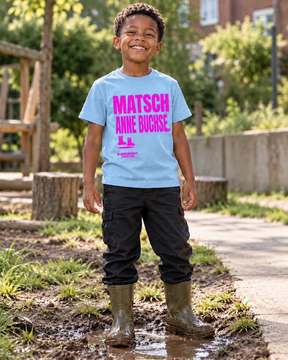 Fiktives Kind im hellblauen T-Shirt mit pinkem Motiv „MATSCH ANNE BUCHSE.“ bei Tageslicht. KI-Produktillustration.
