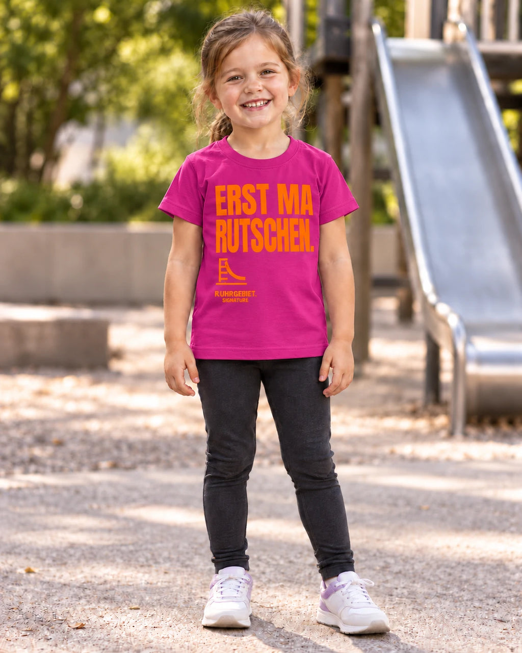 Fiktives Kind im kräftig lilafarbenen T-Shirt mit orangefarbenem Motiv „ERST MA RUTSCHEN.“ auf dem Spielplatz. KI-Produktillustration.