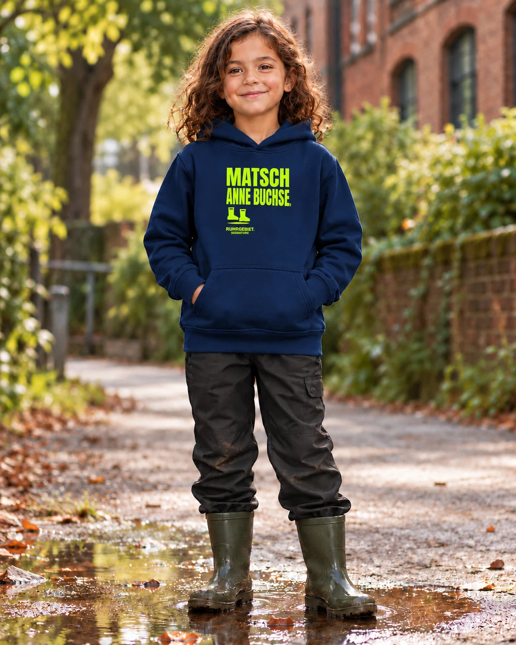 Fiktives Kind im French-Navy-Hoodie mit limefarbenem Motiv „MATSCH ANNE BUCHSE.“ bei Tageslicht. KI-Produktillustration.