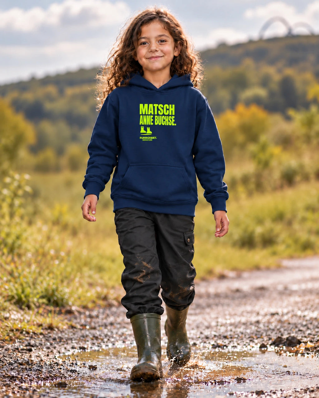 Fiktives Kind im French-Navy-Hoodie mit limefarbenem Motiv „MATSCH ANNE BUCHSE.“ vor Ruhrgebietskulisse bei Tageslicht. KI-Kampagnenbild.